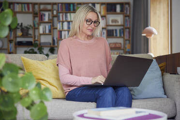 Blond woman using laptop sitting on sofa at home - RBF08880