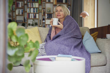 Blond woman wrapped in blanket holding coffee cup on sofa day dreaming at home - RBF08882