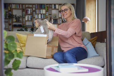Lächelnde Frau, die auf dem Sofa zu Hause die Kleidung im Paket überprüft - RBF08883