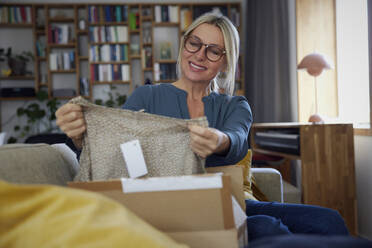 Lächelnde reife Frau, die auf dem Sofa zu Hause die Kleidung im Paket überprüft - RBF08885