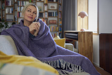 Mature woman wrapped in blanket sitting on sofa day dreaming at home - RBF08886