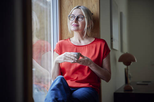 Lächelnde Frau mit Kaffeetasse, die zu Hause am Fenster sitzt - RBF08889