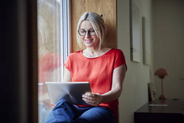 Lächelnde Frau mit Tablet-PC, die zu Hause am Fenster sitzt - RBF08891