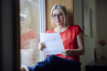 Mature woman wearing eyeglasses reading letter sitting by window at home - RBF08893