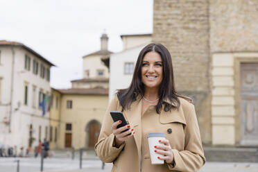 Smiling woman with coffee cup and mobile phone standing in front of building - EIF03938