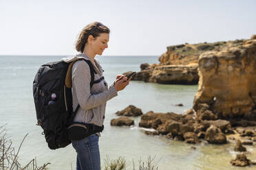 Lächelnde Frau mit Rucksack und Smartphone vor dem Meer stehend - DIGF17823