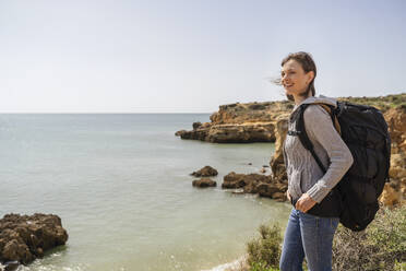 Glückliche Frau mit Rucksack steht vor dem Meer am sonnigen Tag - DIGF17827