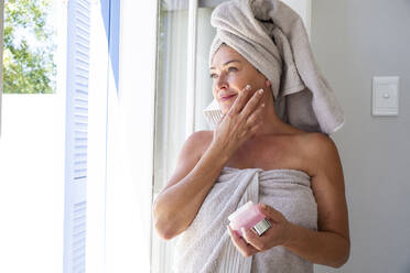 Smiling woman applying moisturizer on face standing by window at home - ESTF00022