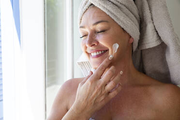 Happy woman with eyes closed applying moisturizer on face at home - ESTF00023