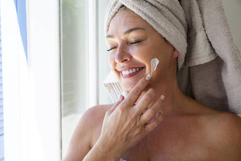 Happy woman with eyes closed applying moisturizer on face at home - ESTF00023