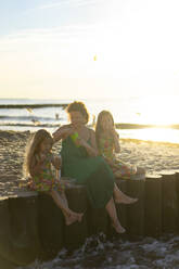 Mother with daughters eating food sitting on wooden post at beach - SSGF00785