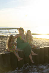Mutter mit Töchtern beim Essen auf einem Holzpfosten am Strand sitzend - SSGF00785