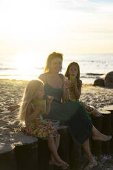 Lächelnde Mutter mit Töchtern beim Essen auf einem Holzpfosten am Strand sitzend - SSGF00786