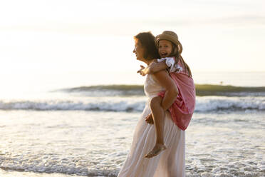Smiling mother giving piggyback ride to cheerful daughter at beach - SSGF00808