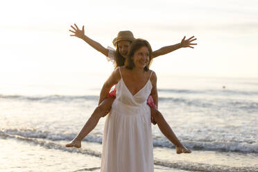 Happy woman giving piggyback ride to daughter enjoying at beach - SSGF00809