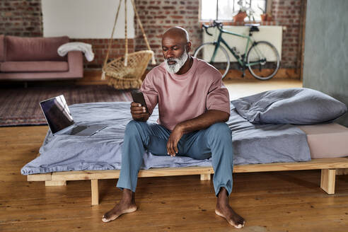 Bald man using smart phone sitting by laptop on bed at home - FMKF07395