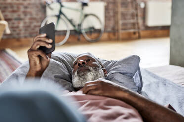 Mann mit weißem Bart, der ein Smartphone benutzt und zu Hause auf dem Bett liegt - FMKF07398