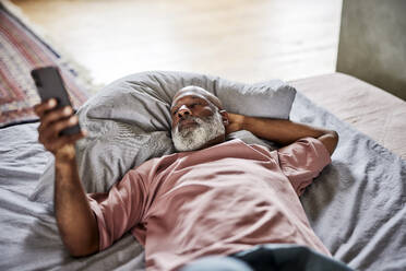 Kahlköpfiger Mann mit Hand hinter dem Kopf, der ein Smartphone benutzt und zu Hause auf dem Bett liegt - FMKF07401