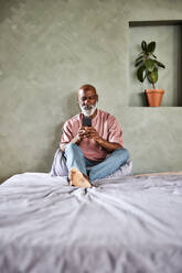 Glatzköpfiger Mann, der zu Hause auf dem Bett sitzend eine SMS über sein Mobiltelefon verschickt - FMKF07403