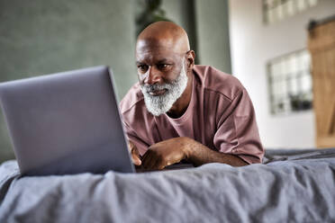Glatzköpfiger Mann mit Laptop auf dem Bett liegend zu Hause - FMKF07406