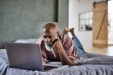 Älterer Mann mit weißem Bart, der einen Laptop benutzt und zu Hause auf dem Bett liegt - FMKF07407