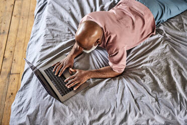 Mann mit Glatze tippt auf einem Laptop, der zu Hause auf dem Bett liegt - FMKF07408