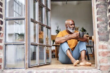 Nachdenklicher Mann mit Glatze und Kaffeetasse am Fenster sitzend - FMKF07424