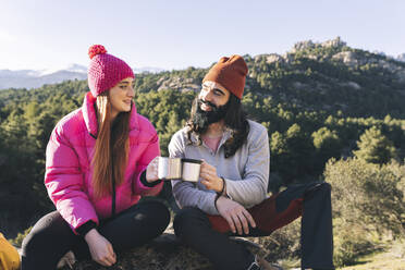 Smiling man and woman toasting mugs sitting on rock - JCCMF06110