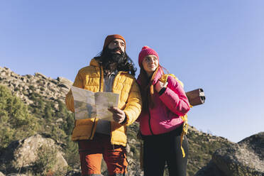 Bearded man with map standing by girlfriend wearing backpack on sunny day - JCCMF06119