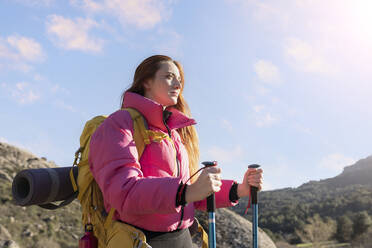 Young woman wearing backpack standing with hiking poles on sunny day - JCCMF06123