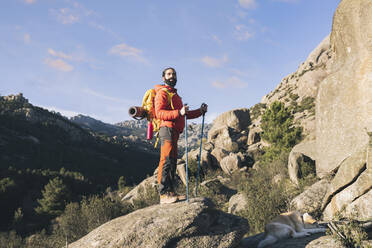 Man wearing backpack standing with hiking poles on rock - JCCMF06125