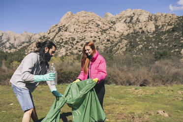 Man and woman with garbage bag collecting plastic bottle on sunny day - JCCMF06130