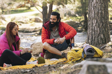 Smiling bearded man looking at girlfriend sitting on picnic blanket in forest - JCCMF06138