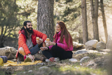 Fröhlicher Mann und Frau mit Essen im Wald sitzend - JCCMF06140
