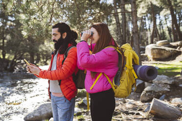 Mann, der das Smartphone seiner Freundin benutzt und durch ein Fernglas in den Wald schaut - JCCMF06154