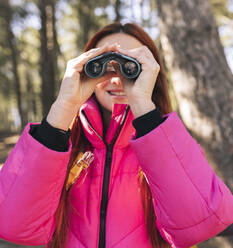 Lächelnde junge Frau schaut durch ein Fernglas im Wald - JCCMF06157