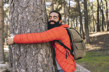 Happy bearded man with eyes closed hugging tree trunk in forest - JCCMF06164