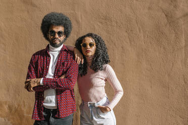 Couple wearing sunglasses standing in front of brown wall on sunny day - MMPF00016