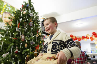 Lächelnder Junge hält Geschenk neben dem Weihnachtsbaum - CAVF96259