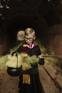 Boy holding magic cauldron with yellow smoke wearing costume in tunnel - GMLF01304