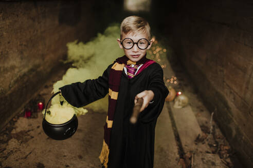 Smiling boy with magic cauldron and magic wand wearing costume standing in tunnel - GMLF01305