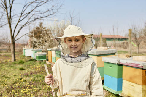 Smiling boy wearing beekeepers protective suit standing in front of containers - ZEDF04535