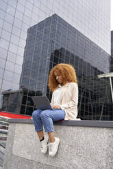 Businesswoman using laptop sitting on wall outside office building - VEGF05585