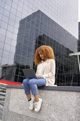 Geschäftsfrau mit Laptop an der Wand sitzend vor einem Bürogebäude - VEGF05585