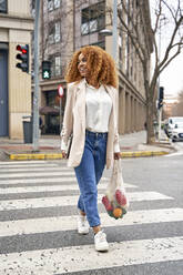 Lächelnde Geschäftsfrau beim Überqueren der Straße mit Obst in einer Netztasche - VEGF05593