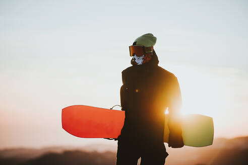 Snowboarder in Sportkleidung und mit Brille hält sein Snowboard bei Sonnenuntergang - CAVF96295