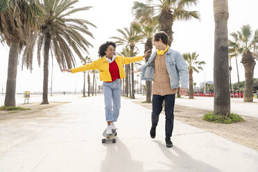 Glücklicher Mann mit Frau beim Skateboardfahren vor Palmen auf dem Fußweg - OIPF01588