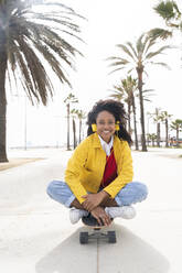 Happy woman wearing headphones sitting on skateboard in front of palm trees - OIPF01591