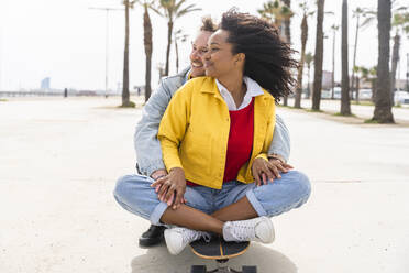 Happy couple sitting on skateboard at footpath - OIPF01592