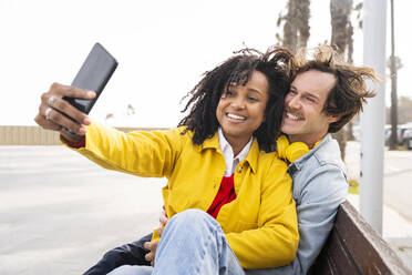 Lächelnde Frau mit Mann, der ein Selfie mit dem Handy macht, auf einer Holzbank sitzend - OIPF01620